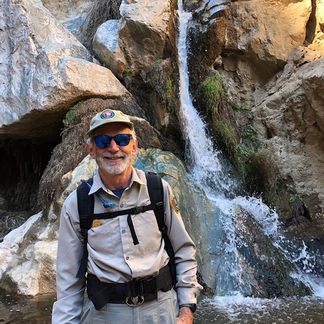 Volunteer Dave in uniform standing in front of rocky waterfall, smiling for a picture. 