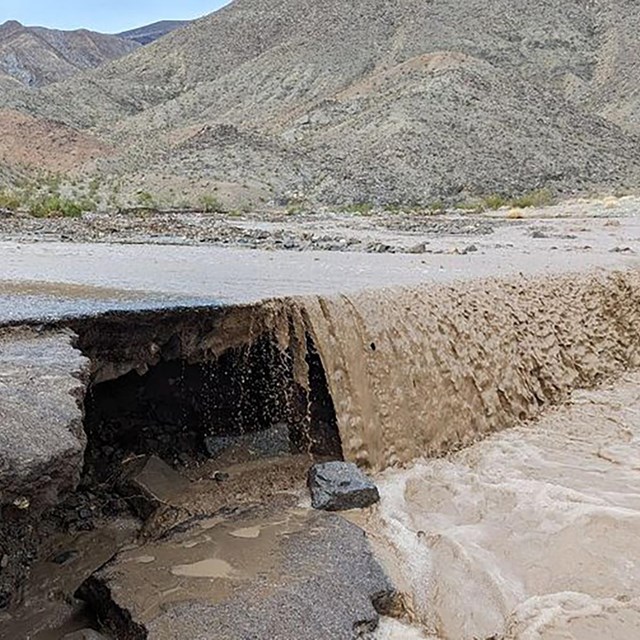 Muddy water pours over the side of broken pavement on a road. Mountains line the background.