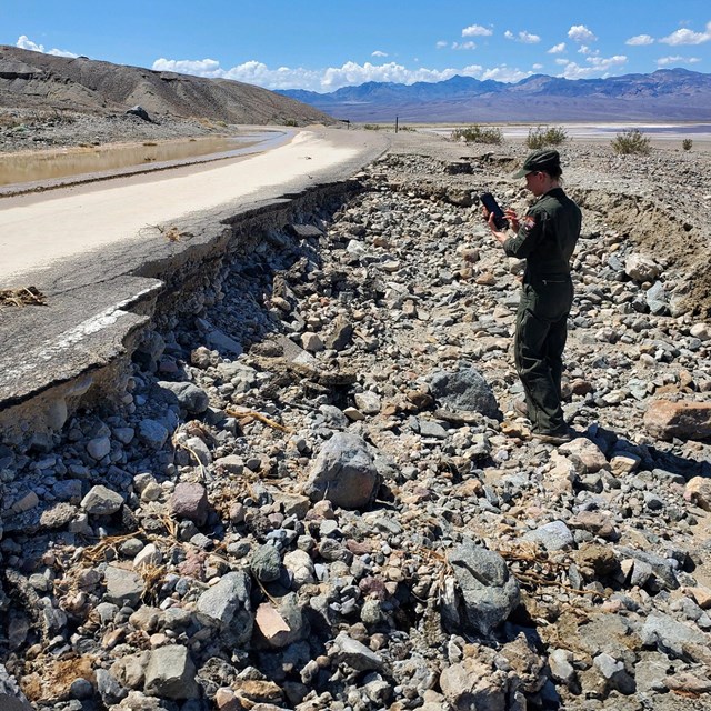 Uniformed ranger stands on rock and debris below the undercut section of a paved road.