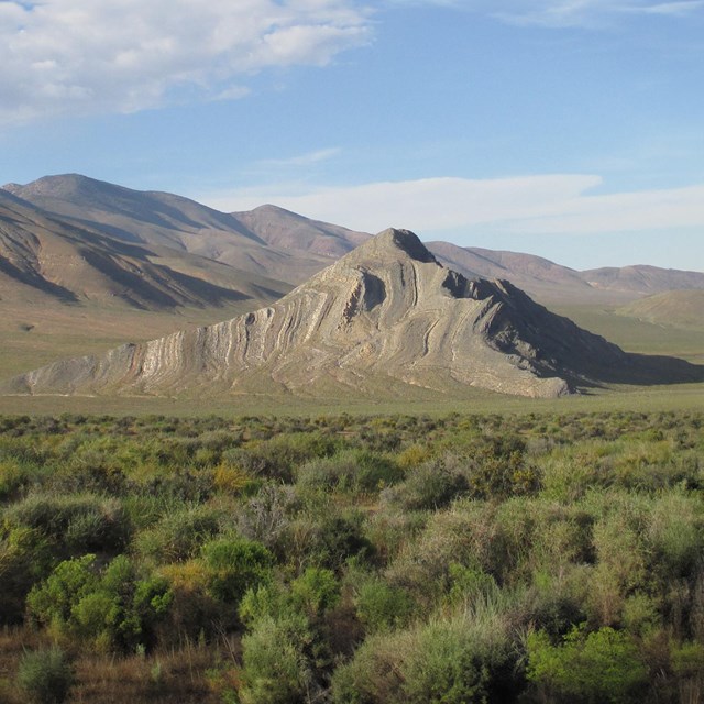 A striped butte stands in the middle of a green valley with shrubs in the foreground.