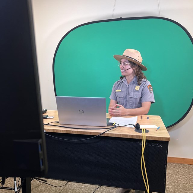 Uniformed ranger stands in front of green screen behind table.
