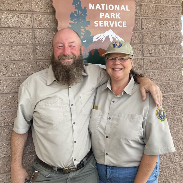 Volunteers Greg and Lisa in uniform, standing in front of a National Park Service Arrowhead sign.