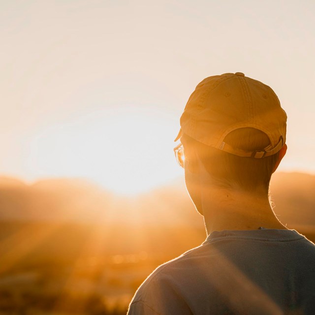 An individual in a t-shirt and ball cap watches the sunset over the distant mountains as light beams