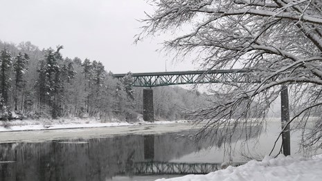 A green bridge spans a river surrounded by a landscape dusted with snow. 