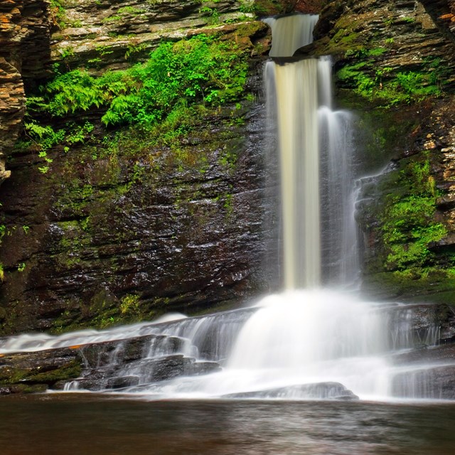 A waterfall flows over moss covered rocks into a pool below.