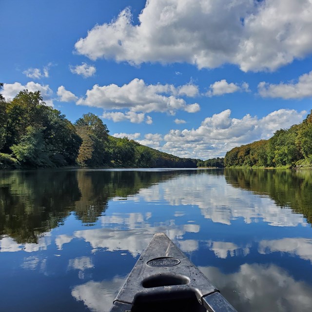 The front end of a canoe pointing downriver with a blue sky and green trees.