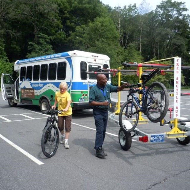 A shuttle bus with a trailer attached to it. People are loading bicycles onto the trailer.