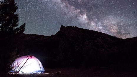 A glowing tent sitting beside a tree. In the background is a sharp ridgeline beneath a starry sky.