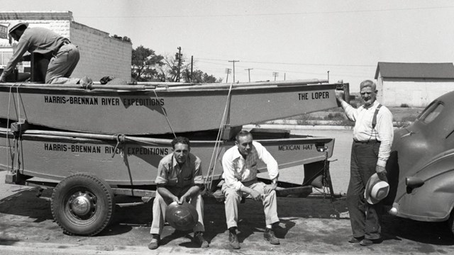 A black and white photo of three men beside a truck hauling two boats with "loper" printed on them.