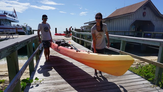 2 people standing on a wooden dock, carrying a kayak
