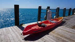A long orange and yellow kayak vessel resting on a wooden dock beside a blue ocean and blue sky