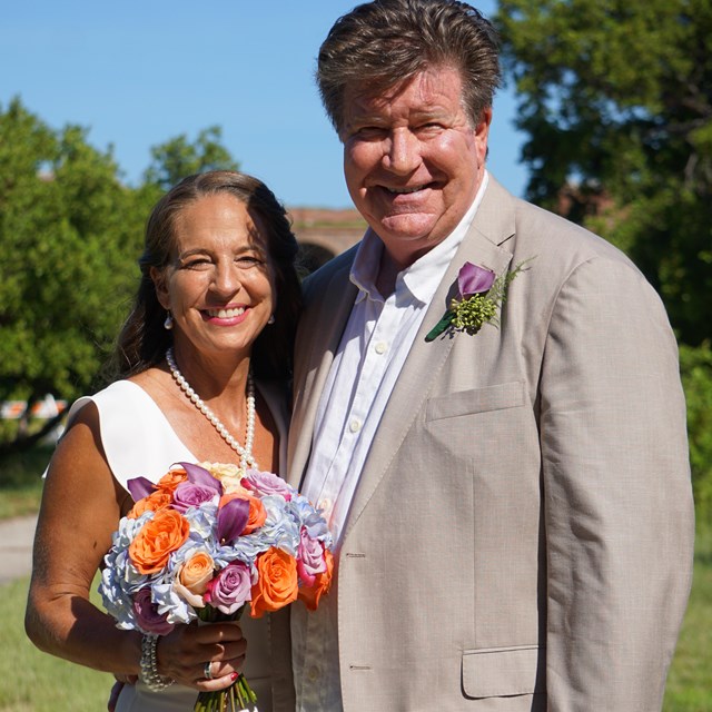 two people standing and smiling with trees and a fort structure in the background