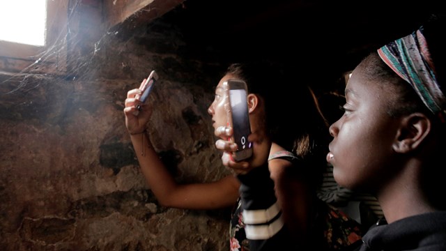 Color photo of two teen girls taking photos of spider webs in the basement window.