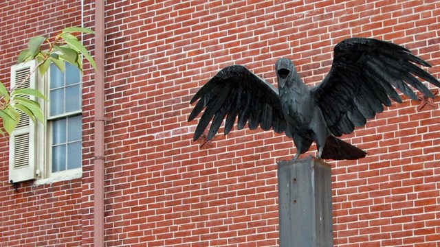 Color photo showing a statue of a raven with wings outspread  in front of a brick building.