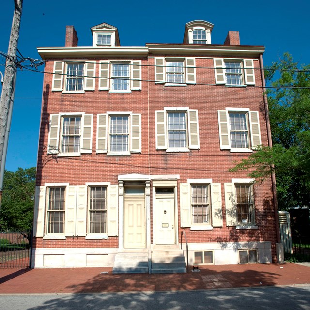 Color photo of the exterior of two red brick row homes, each with three floors.