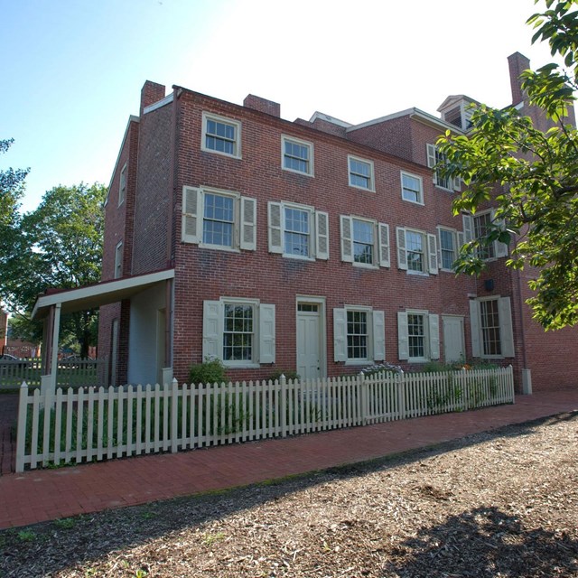 Color photo of three story red brick home behind a wooden fence.