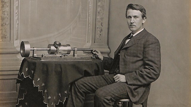 Younger Thomas Edison sitting at ornate table with original phonograph atop it.