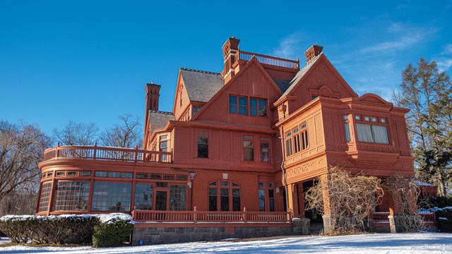 A three story brick red mansion on a lawn surrounded by trees.