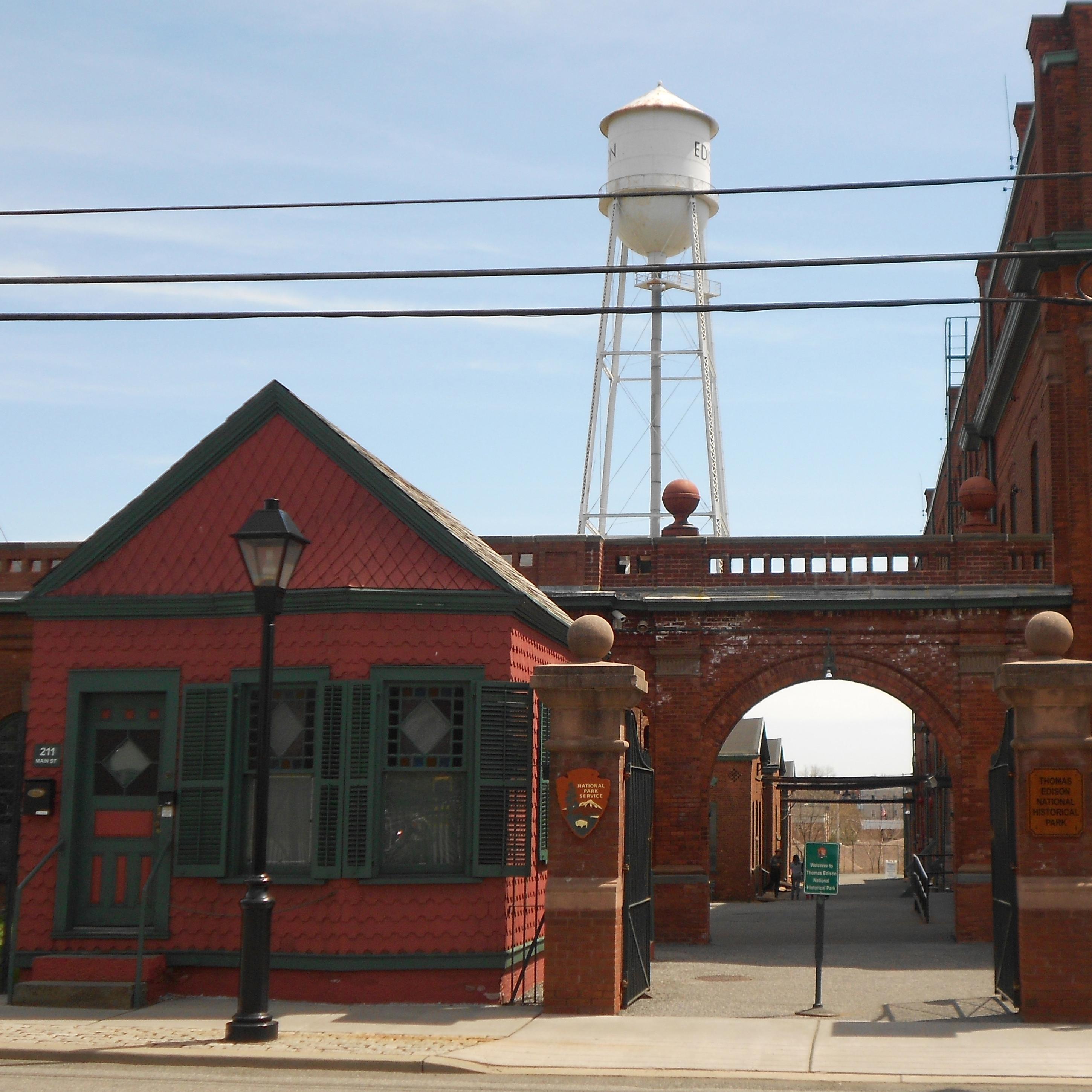 Entrance has small red wooden building next to large brick building with water tower in background.
