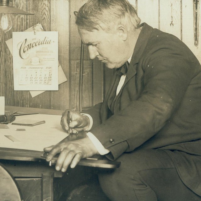 Thomas Edison sitting at table writing on paper.