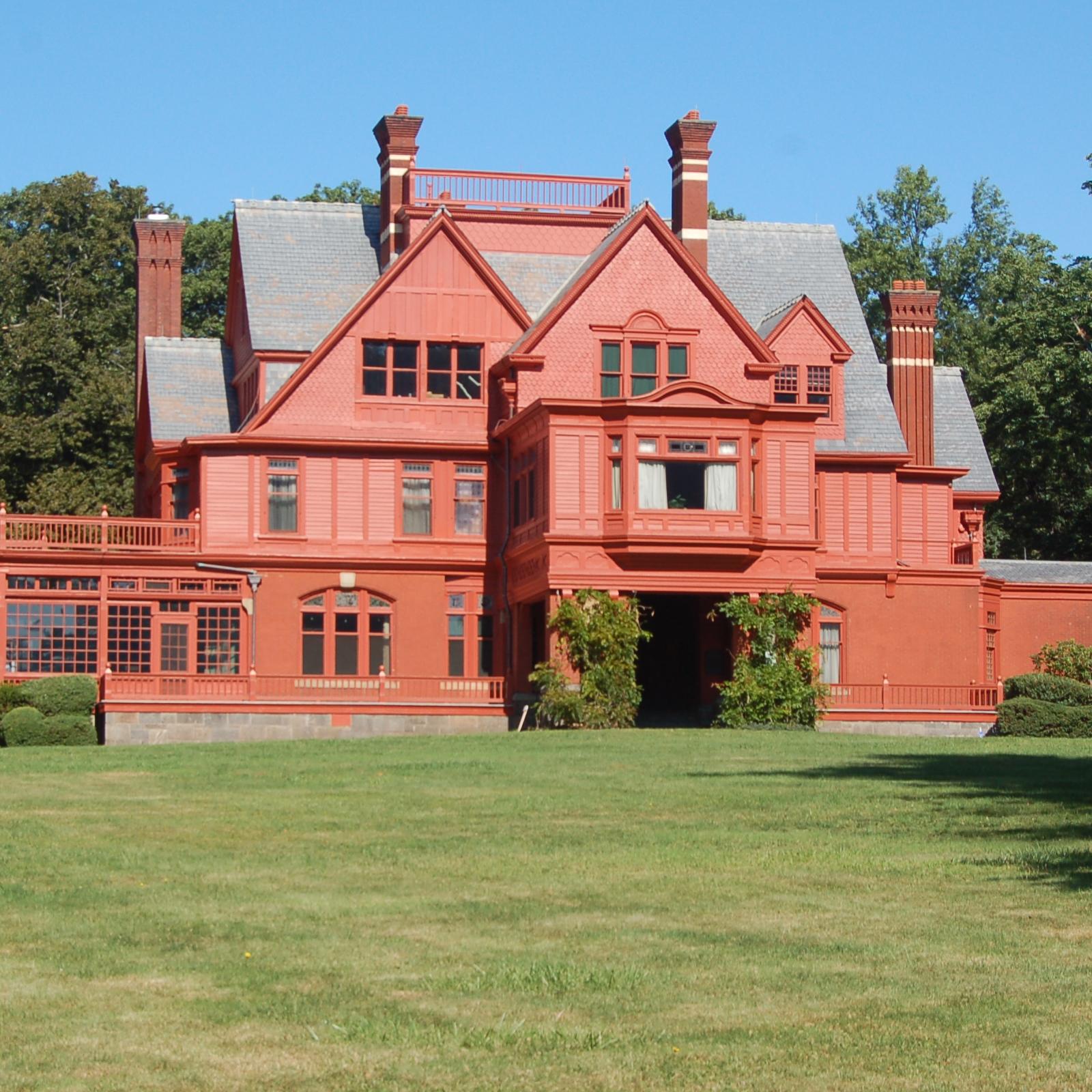 A three story brick red mansion on a lawn surrounded by trees.