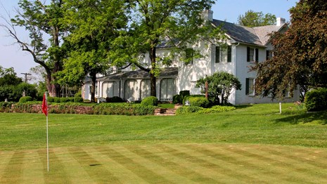 A photo of a large white house partially screened by trees and golf putting green in the foreground.