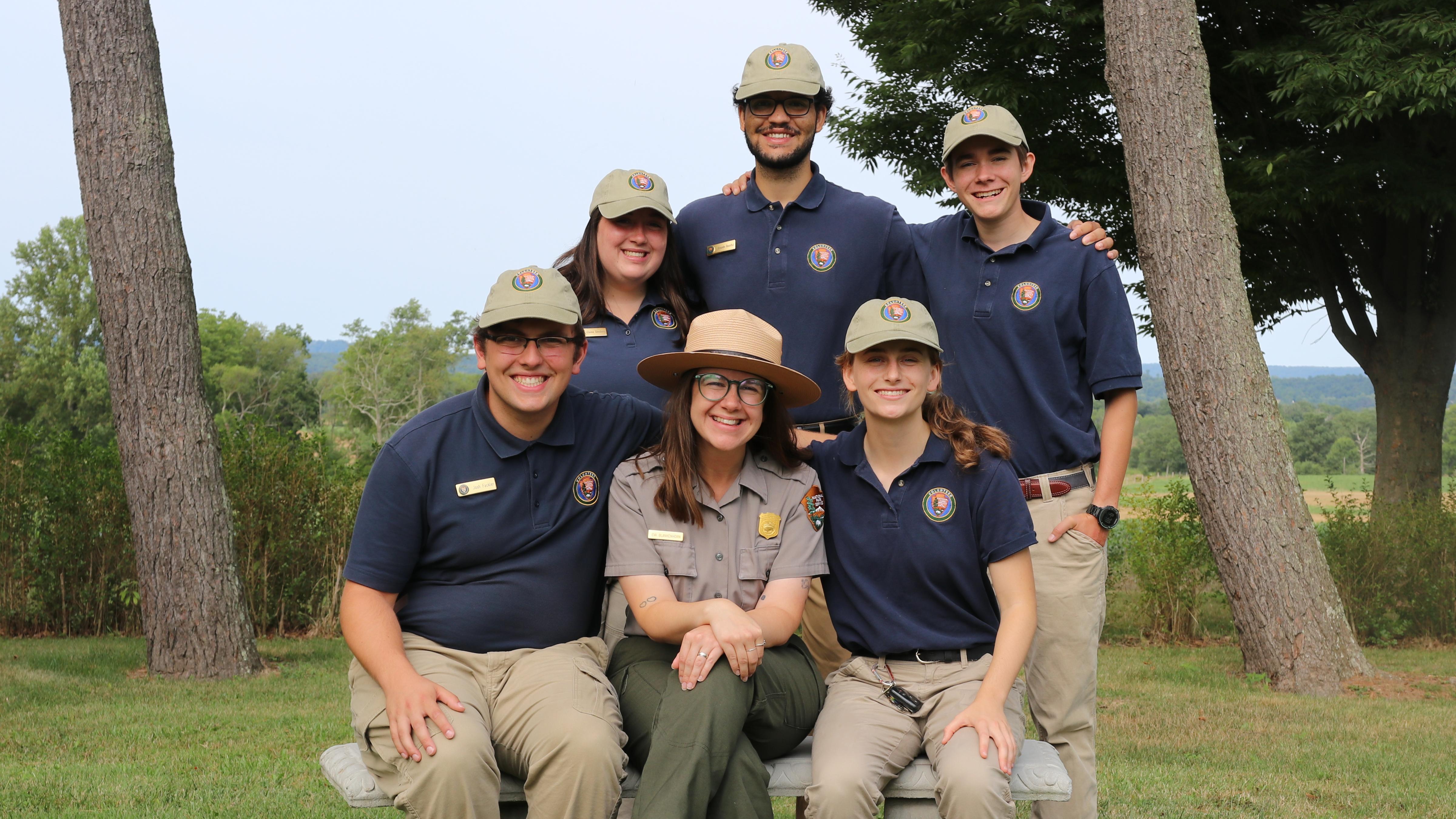 A group of five interns in blue polo shirts and khaki hats stand around a park ranger
