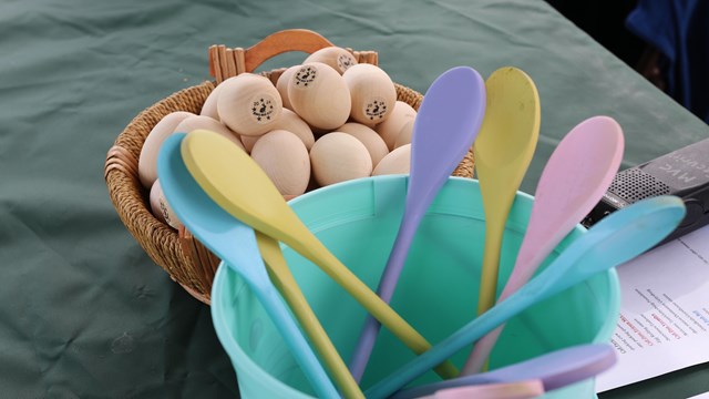 A basket of wooden eggs sits next to a bucket of pastel pained wooden spoons