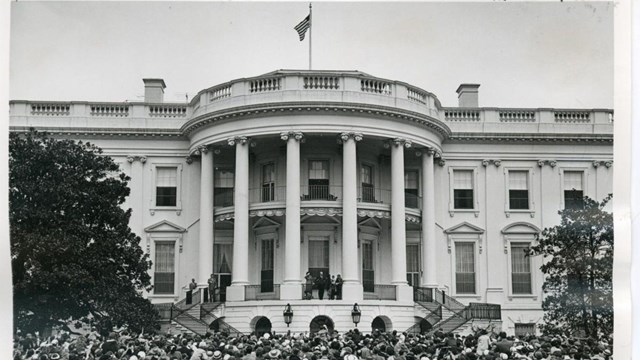 A black and white photograph of a large crowd gathered in front of a large two story white building