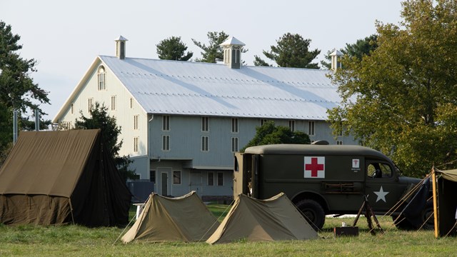 A color image of a WWII vehicle and tents with a green bank barn in the background.