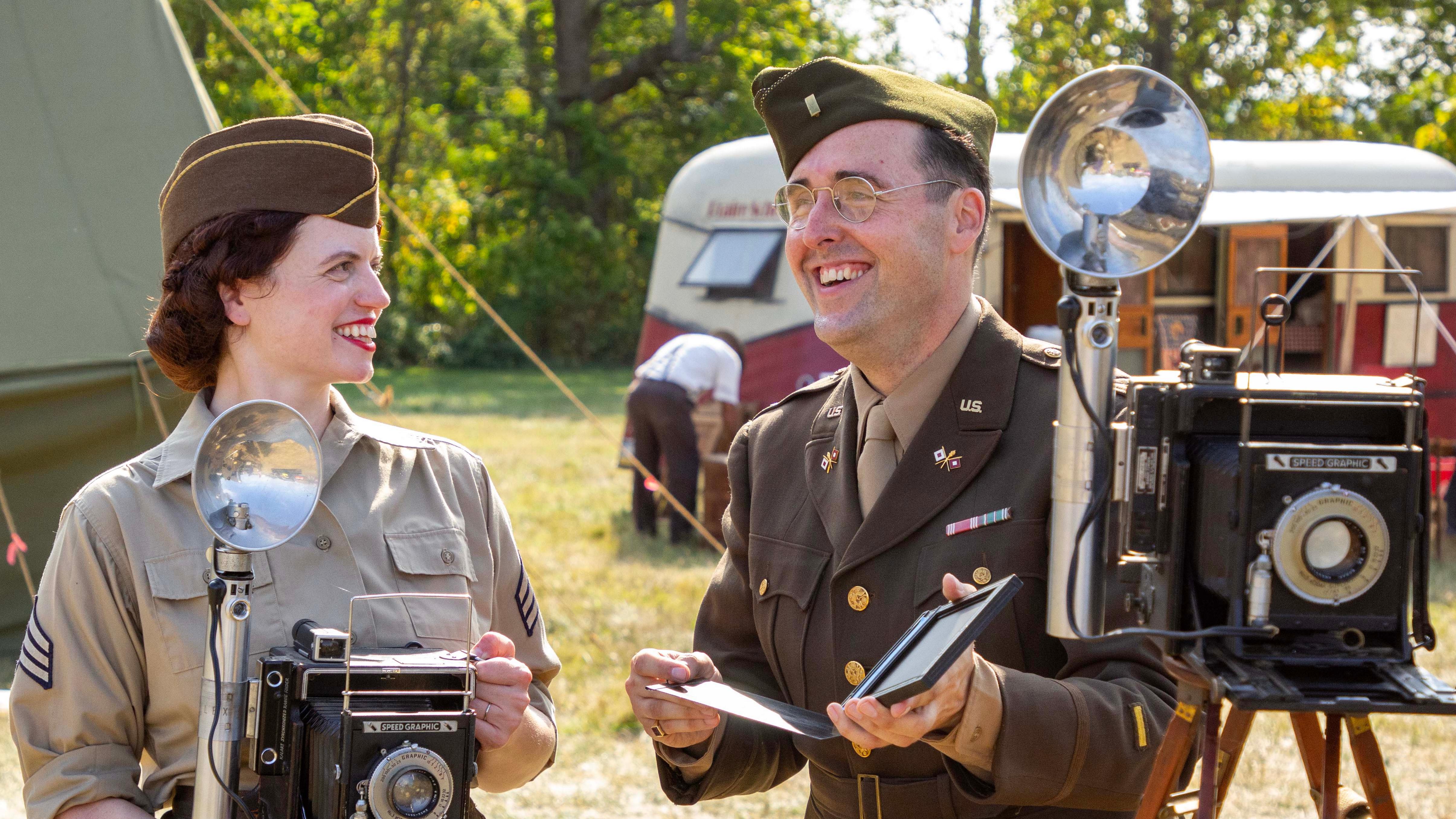 Two volunteers in WWII uniforms smile and pose holding historic cameras