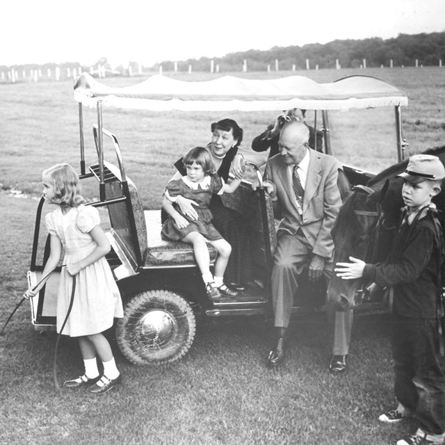 A black and white photo of two adults, three children, a horse, and a four passenger golf cart.