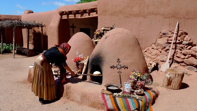 A woman uses a wooden tool to remove food from an adobe earthen oven, set against an adobe building.