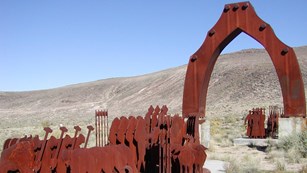 A statue of soldiers meeting travelers under an arch, set in the desert.