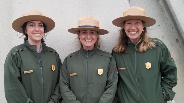 Three park rangers with straw hats stand in front of a wall.