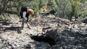 A hiker with a backpack stoops down to look at a hole in the cooled lava. 