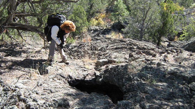 A hiker with a backpack stoops down to look at a hole in the cooled lava. 