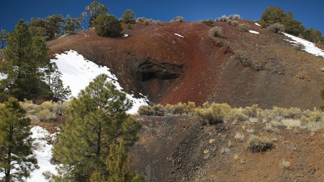 A reddish hill with pine trees and snowy patches.