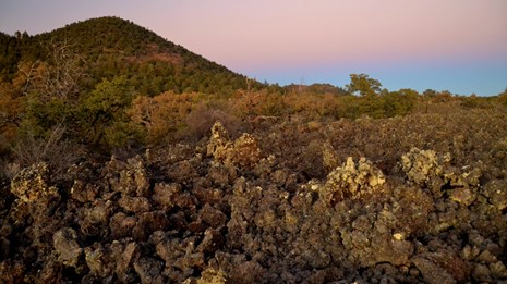 A rubbly lava flow with a volcanic hill in the background at sunset.