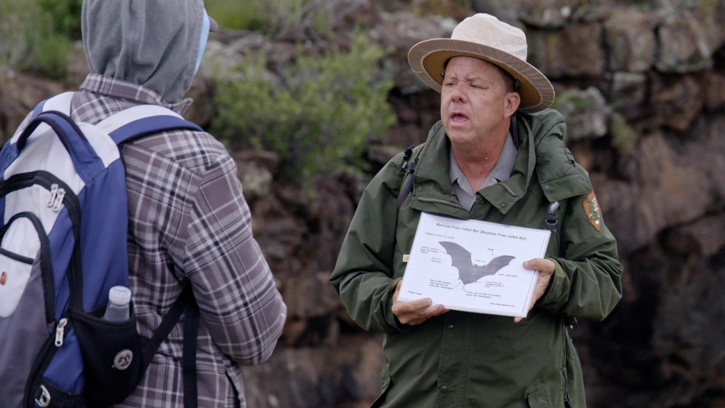 A park ranger holding an image of a bat with two people watching him.