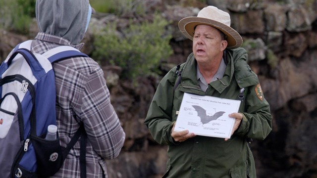 A park ranger holding an image of a bat with two people watching him.