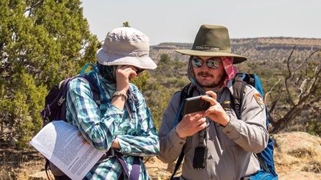 A park ranger in hiking gear and sun protection shows a hiker something on his phone.