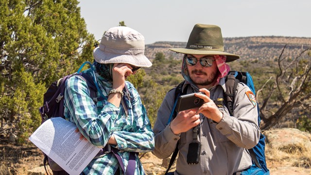 A park ranger in hiking gear and sun protection shows a hiker something on his phone.
