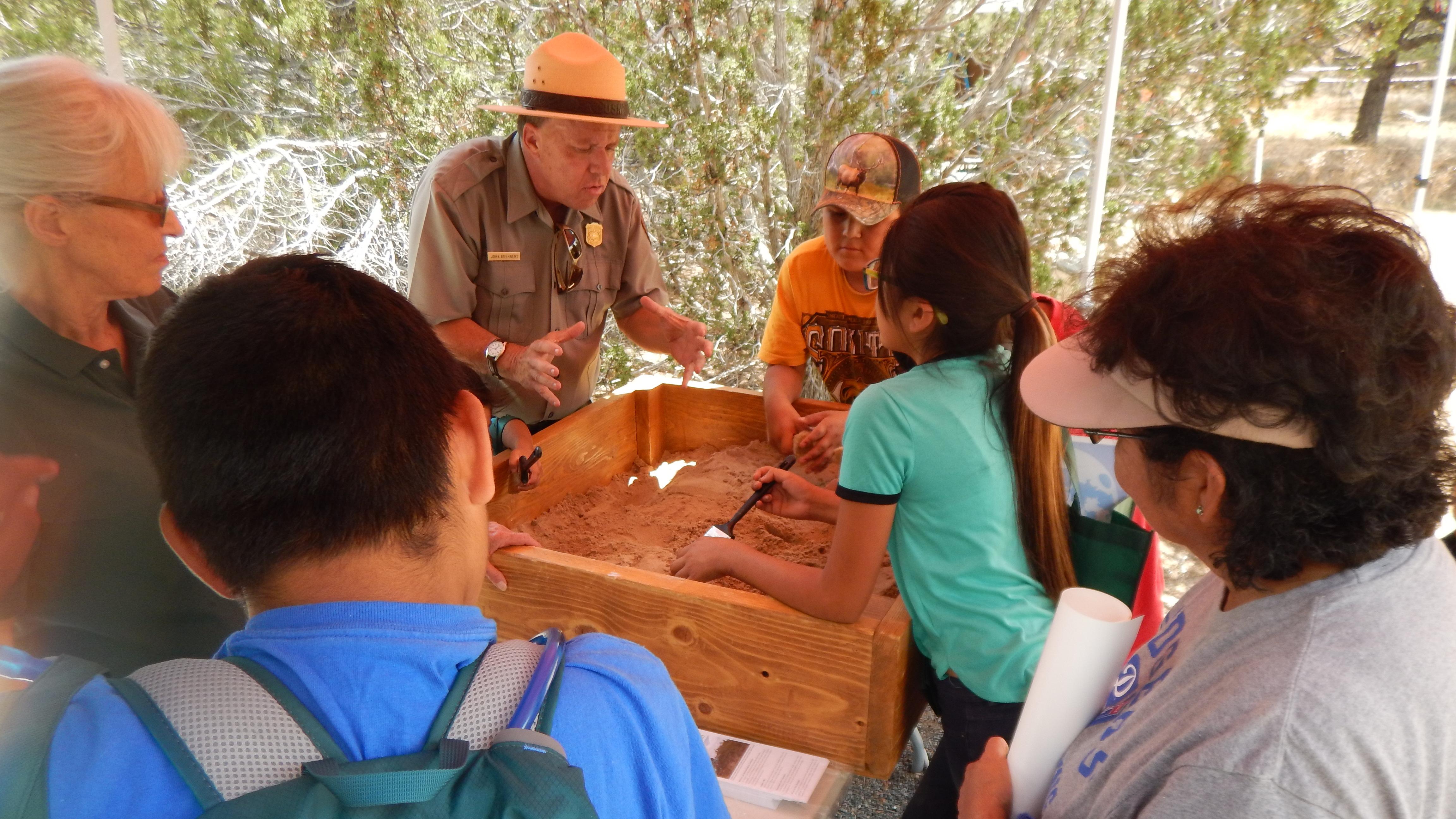 A ranger presents to a group of students at a table. Everyone is looking into a box of sand.