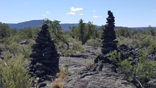 Two person-sized piles of rock tower above shrub-covered rock.