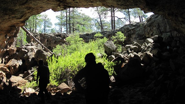 Two cavers are silhouetted as they exit a cave towards a forest above.