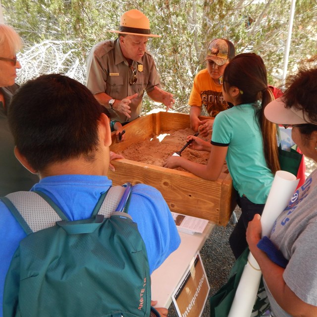 Ranger is presenting to a group a students at a table. Everyone is looking into a box of sand.