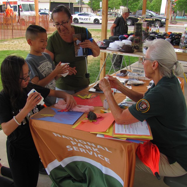 Park volunteer sits at a table helping 2 children and a parent with an arts project.