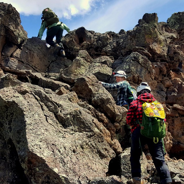 Three people dressed in hard hats climb up a steep hill of boulders.