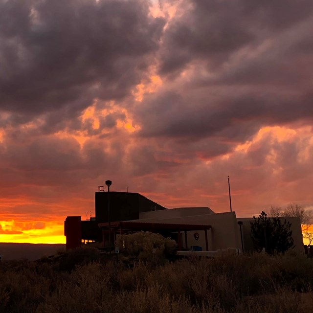 A golden sunset silhouettes an angular stucco structure.  Clouds fan out across the sky.